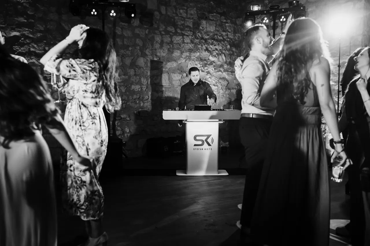 Tanzende Hochzeitsgäste in einem Gewölbe, im Hintergrund der DJ am Pult, schwarzweiß